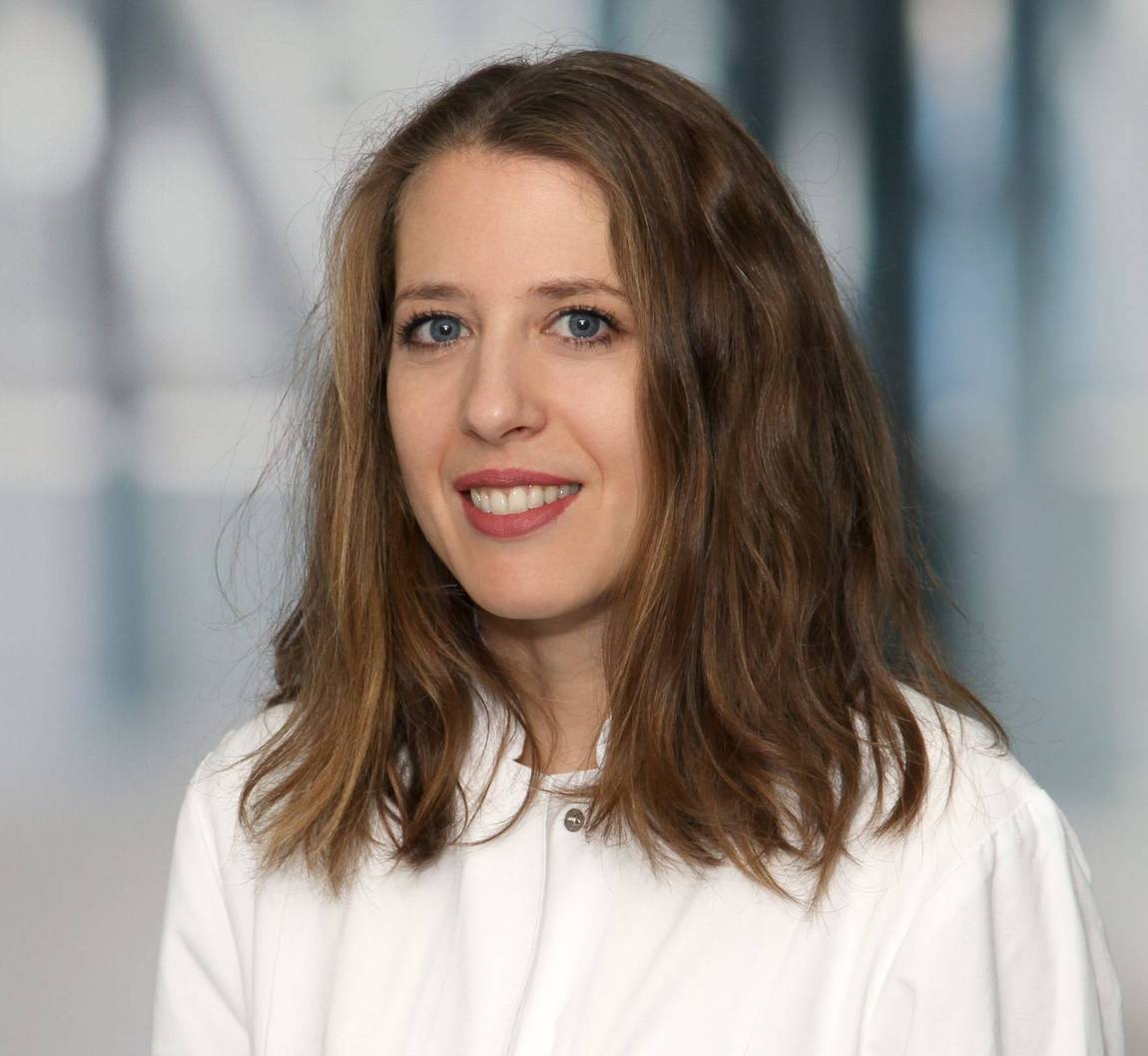 Eine Frau mit gewelltem, schulterlangem braunem Haar und blauen Augen lächelt in die Kamera. Sie trägt ein weißes Oberteil und der Hintergrund ist mit kühlen Tönen sanft verschwommen.